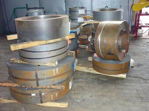 Skladowane kręgi blachy stalowej S355MC na drewnianych paletach w hali produkcyjnej, widoczne napisy na kręgach.